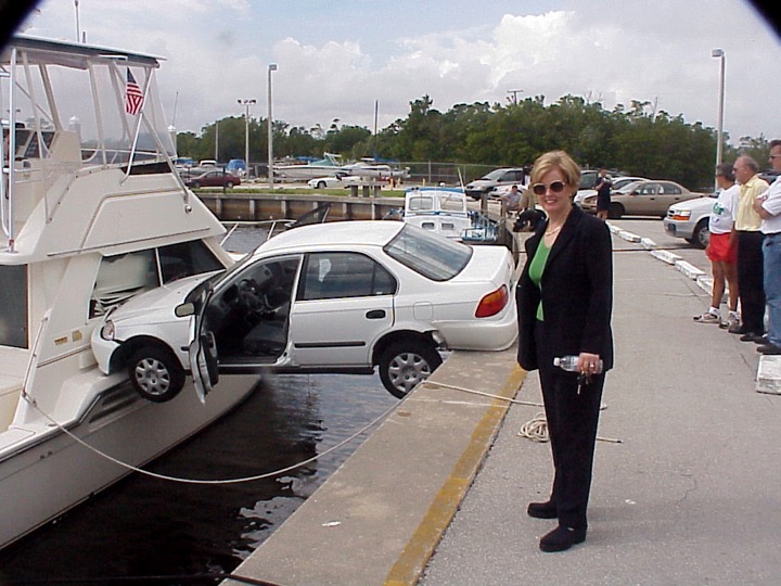 Coche caído entre el espigon de un puerto y un yate, mujer en primer plano